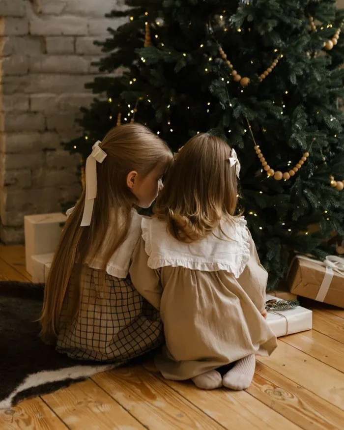 Two little girls sitting together on the floor in front of a tall Christmas tree, seen from behind