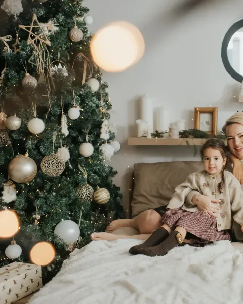 A family of five laughing together on a bed in front of a lit Christmas tree, the youngest on her mother's lap