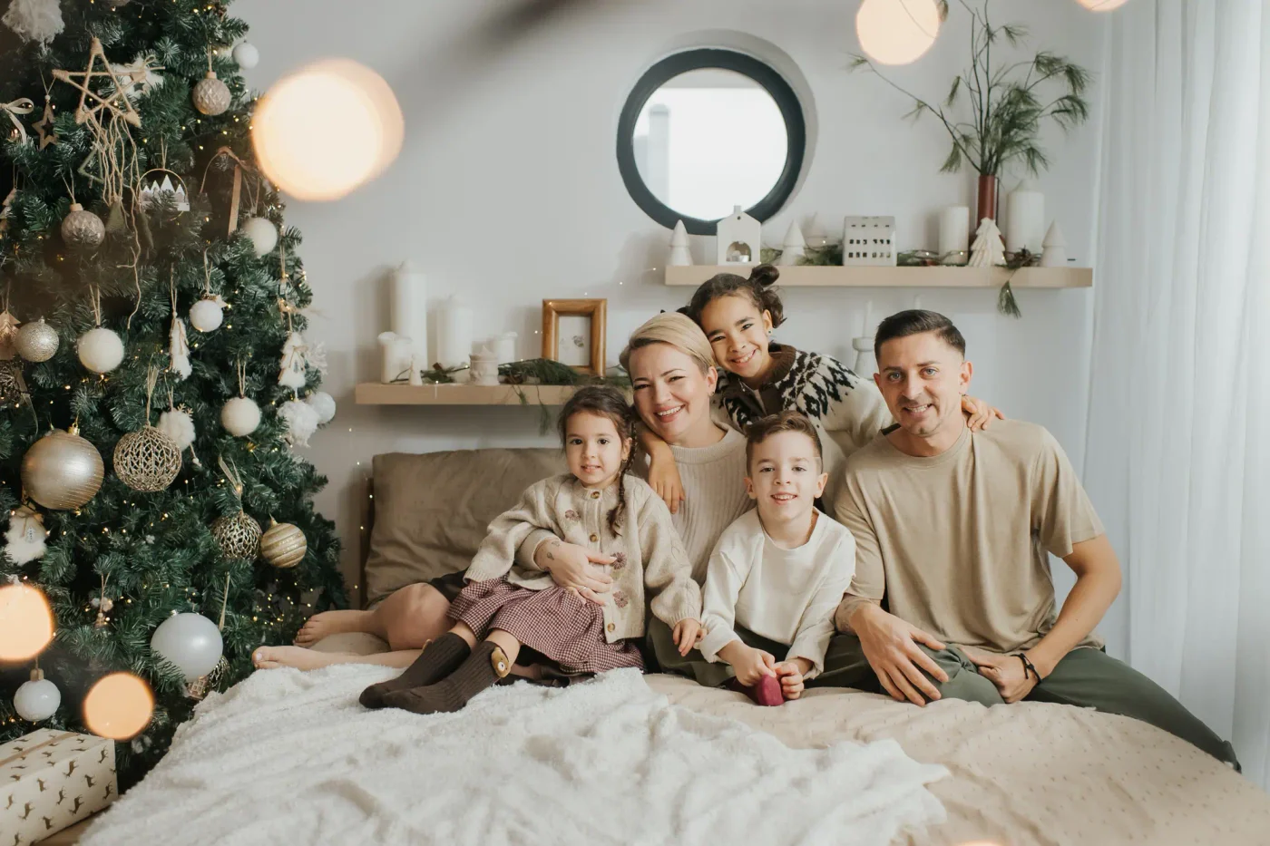 A family of five laughing together on a bed in front of a lit Christmas tree, the youngest on her mother's lap