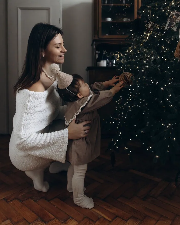 A mother crouching beside her toddler as he reaches for the ornaments on a lit Christmas tree