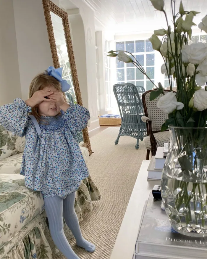 A small girl in a blue floral dress covering her eyes with her hand in a bright bedroom