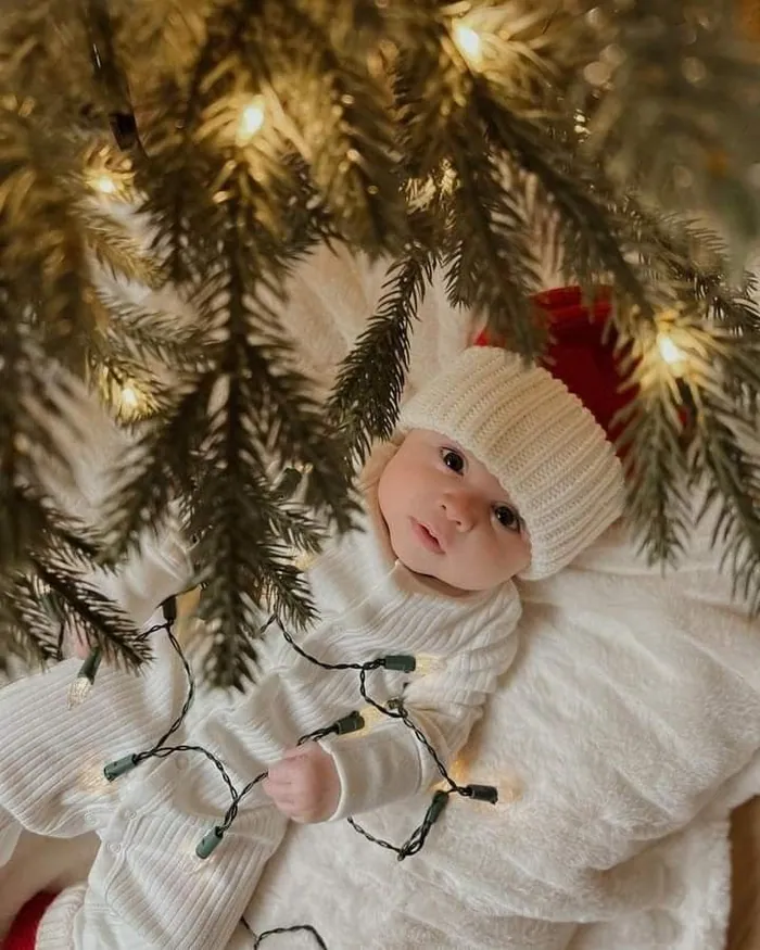A baby in a cream knitted hat lying under the branches of a Christmas tree, a string of lights beside him
