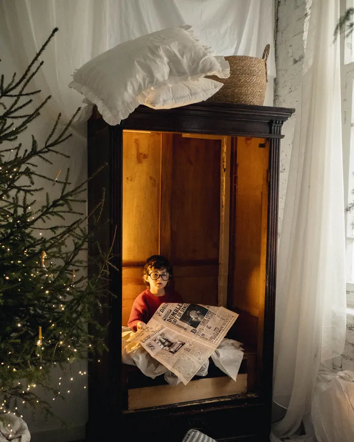 A small boy hiding inside a wooden wardrobe, peeking out over a newspaper