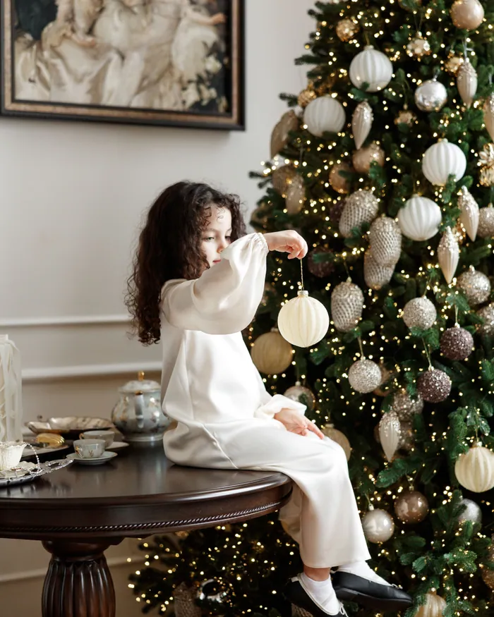 A small girl in a white dress sitting on a round table, reaching towards the ornaments on a tall Christmas tree