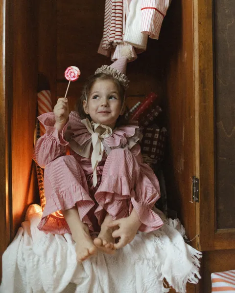 A small girl in pink ruffled pyjamas holding a lollipop, sitting in an open wardrobe hung with stars