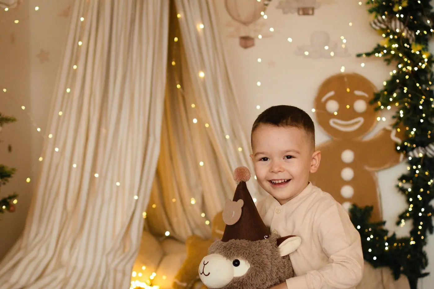 A small boy laughing on a toy rocking reindeer in a children’s room with a canopy, fairy lights and a gingerbread figure