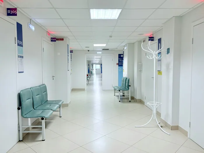 A long, bright hospital corridor with white floors, pale blue waiting chairs and ward signs