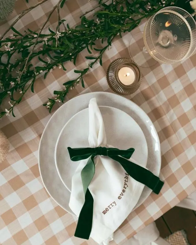 A plate with a white napkin tied in a green velvet bow on a beige gingham tablecloth, a candle beside it