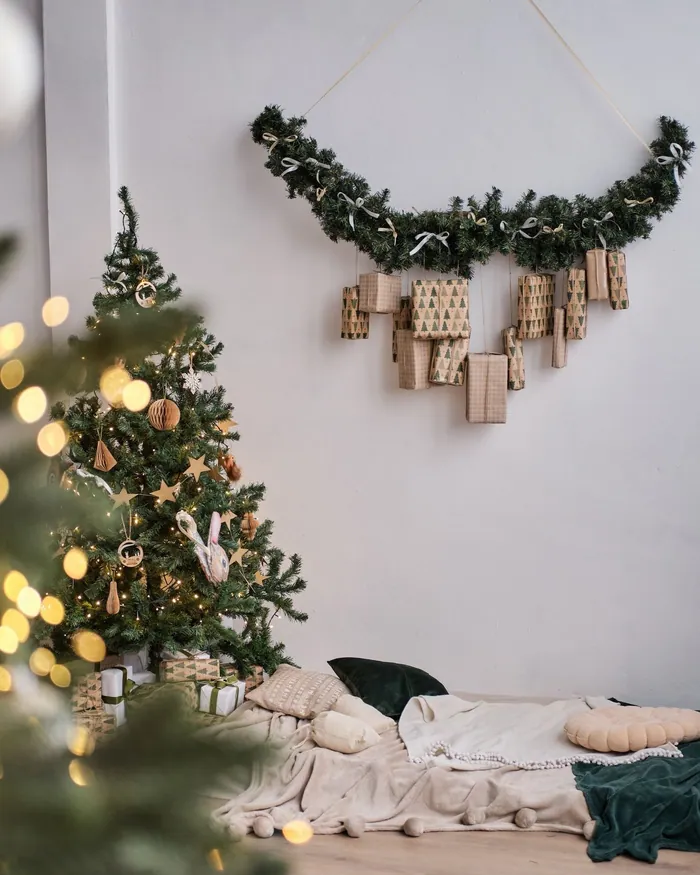 A row of small paper advent bags hung along a fir garland on a white wall, a lit tree beside it
