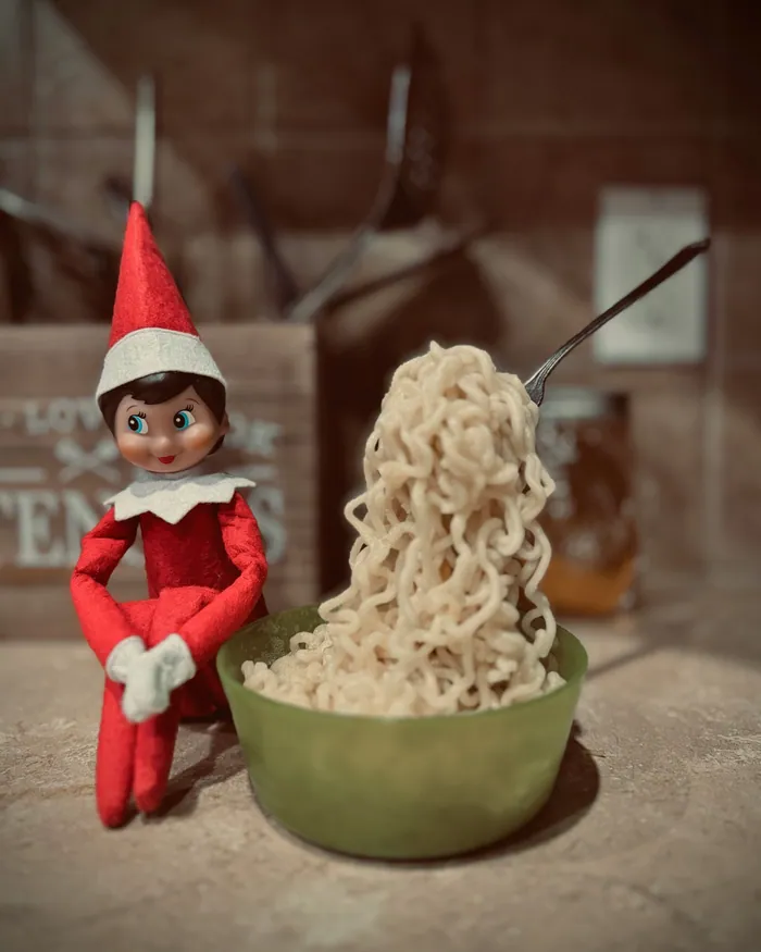 A toy elf sitting on a kitchen counter beside a green bowl of noodles with a fork standing in it