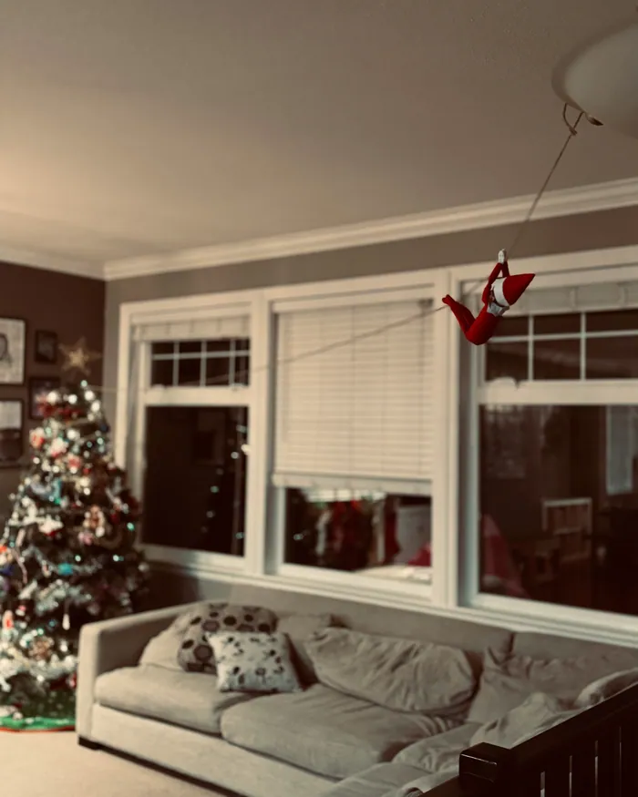 A toy elf hanging from a line strung across a living room, a lit Christmas tree in the corner