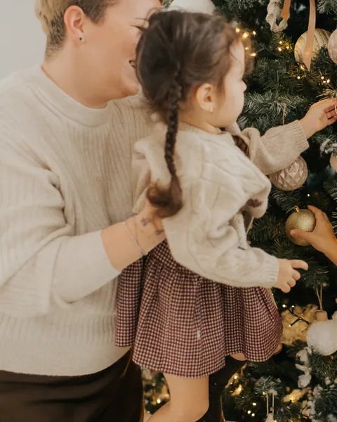 A mother lifting her small daughter in a cream cardigan to hang a bauble on a lit Christmas tree