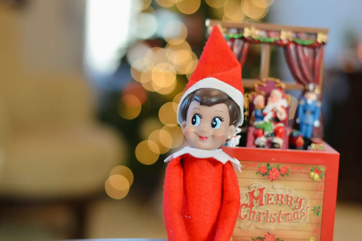 A toy elf in a red suit sitting beside a wooden Merry Christmas box, Christmas lights glowing behind him