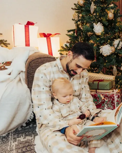 A mother, father and baby in matching beige gingham pyjamas reading a book by the tree, their small white dog in the same pyjamas