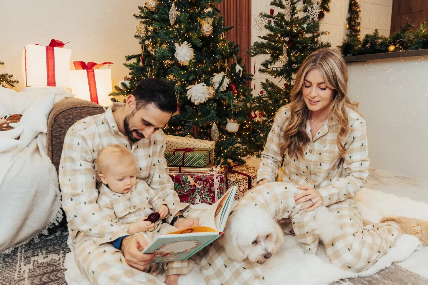 A mother, father and baby in matching beige gingham pyjamas reading a book by the tree, their small white dog in the same pyjamas