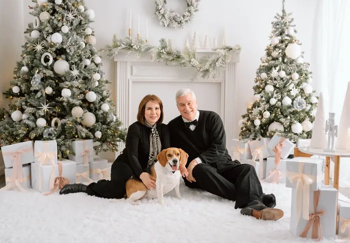 A grandmother and grandfather sitting on a white rug with their beagle between two decorated trees