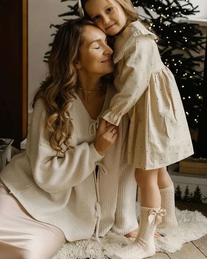 A mother hugging her small daughter in a cream dress in front of a Christmas tree