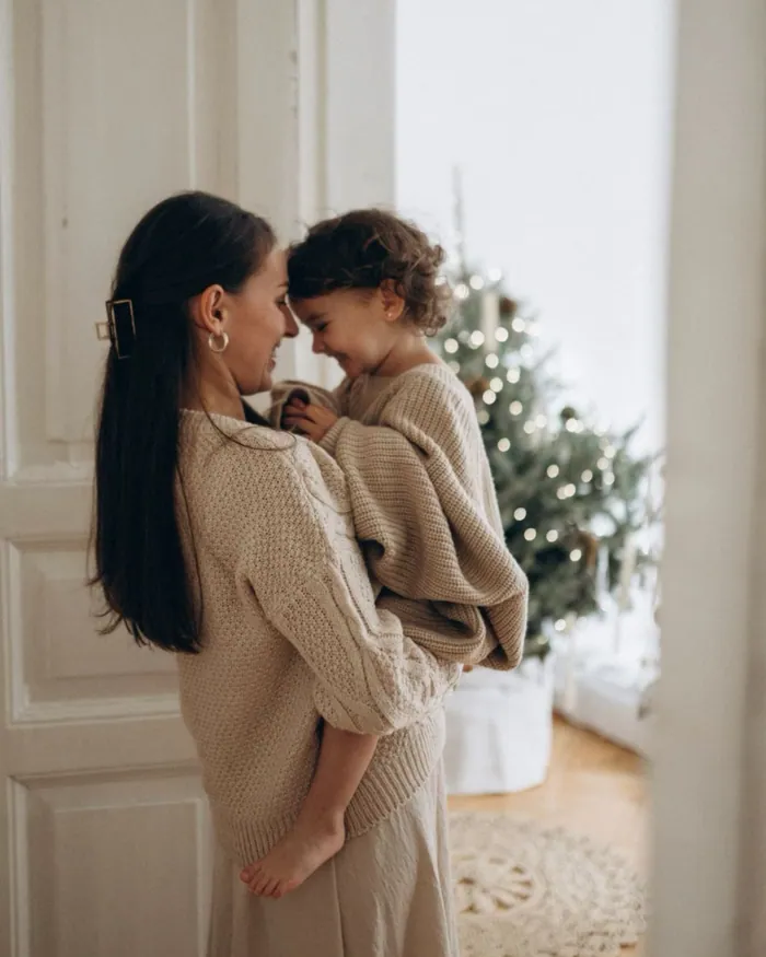 A mother holding her small daughter close in a doorway, the Christmas tree lit behind them