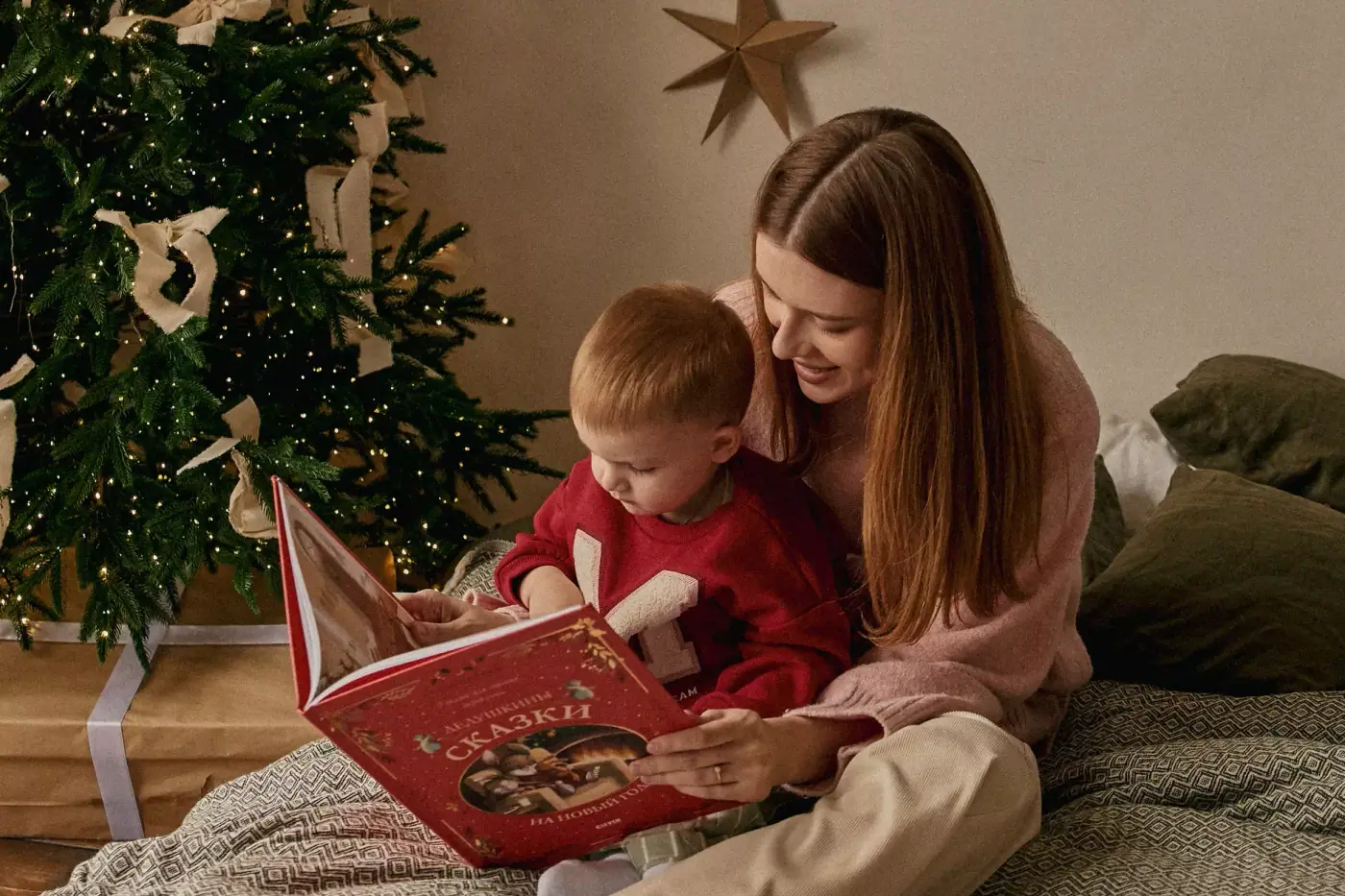 A mother reading a picture book to her small son on a blanket beside a Christmas tree