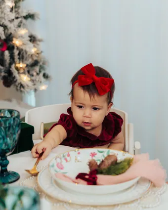 A baby girl in a red velvet bow sitting up at the table in front of a floral plate and a teal goblet
