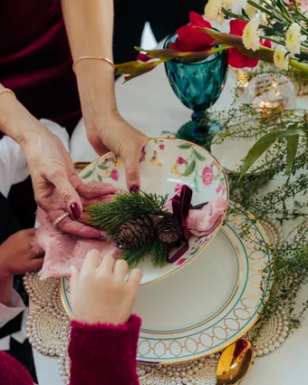 Hands tucking a sprig of pine and pine cones into a pink napkin on a floral plate