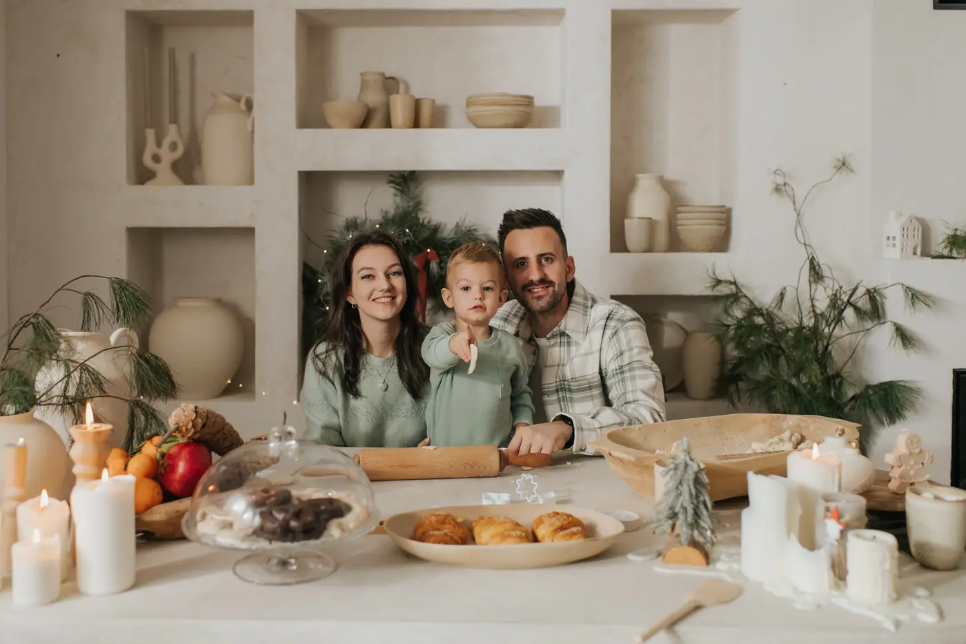 A family of three smiling behind a laid table with pillar candles, a cake dome, croissants and a bowl of fruit, greenery on the shelves behind them