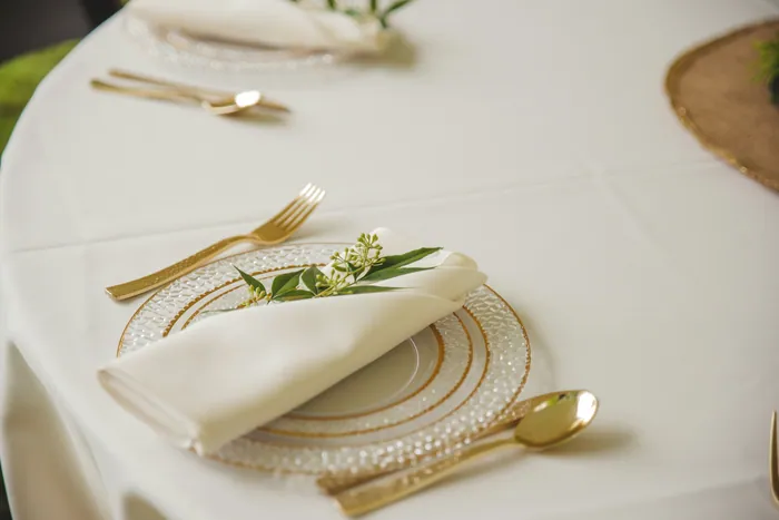 A folded ivory napkin with a sprig of green on a gold-rimmed glass plate with gold cutlery