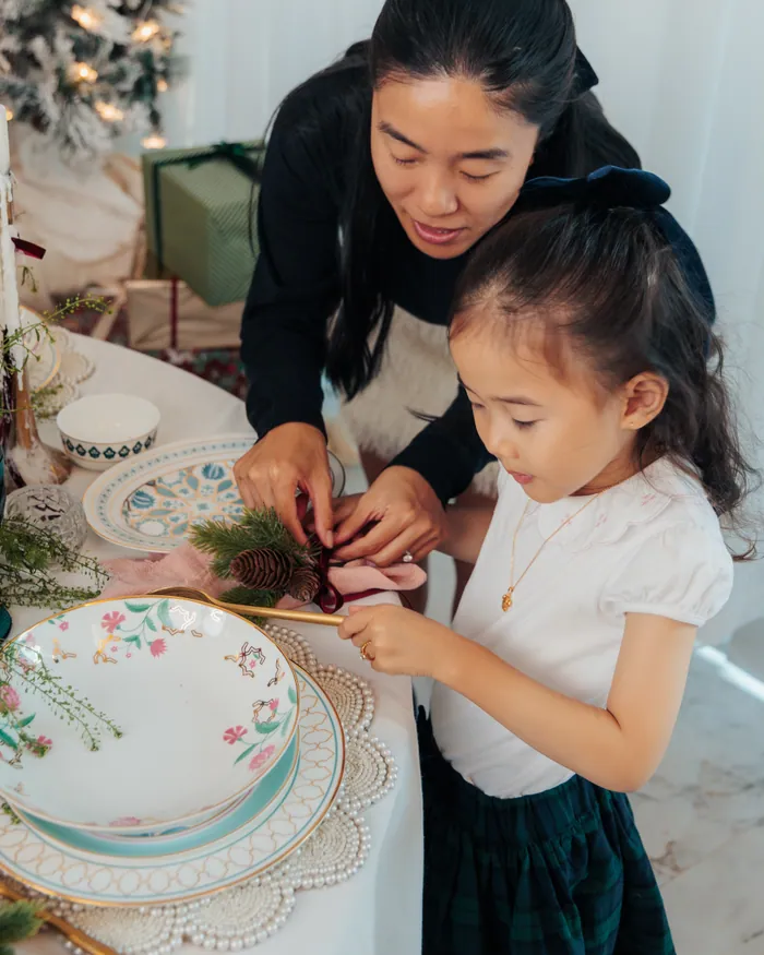 A mother and her young daughter tucking sprigs of pine into pink napkins on floral plates at a laid Christmas table