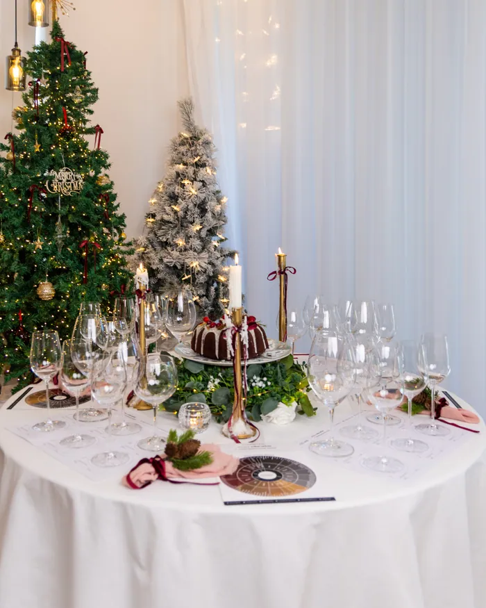 A round table in a white cloth laid with clear glasses, pink napkins tied with velvet, brass candlesticks and a chocolate bundt cake, two lit trees behind