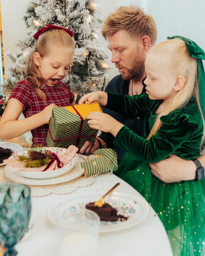 A father and his two daughters opening a wrapped present at a laid table, a chocolate cake and tree lights beside them