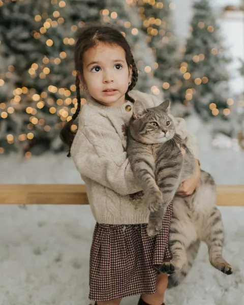 A small girl in a cream cardigan and check skirt holding a grey tabby cat in the snow