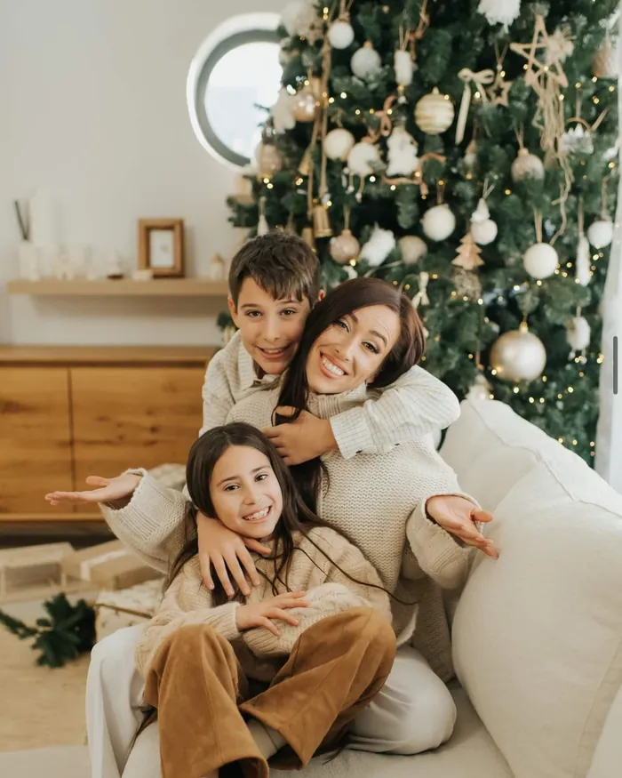 A mother hugging her son and daughter on a white sofa beside a Christmas tree, all three laughing