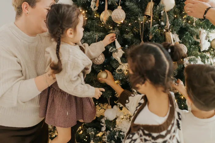 A mother lifting her small daughter to hang a bauble on the tree while two older children reach for ornaments beside her