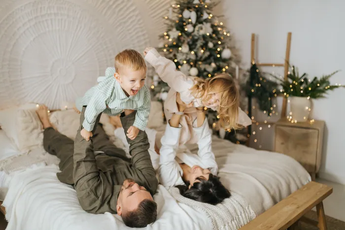 Two parents lying on a bed lifting a laughing toddler and a small girl up into the air, a Christmas tree behind them