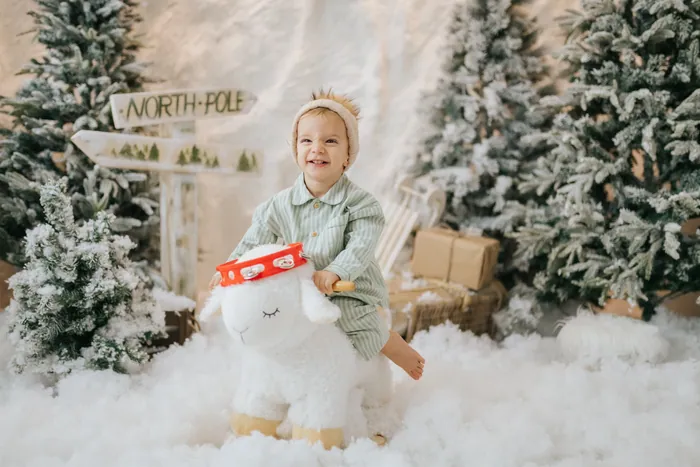A toddler in green striped pyjamas laughing as he rides a toy sheep through the snow, holding a tambourine