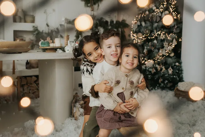 Three siblings hugging and laughing together in the snow, fairy lights glowing out of focus around them