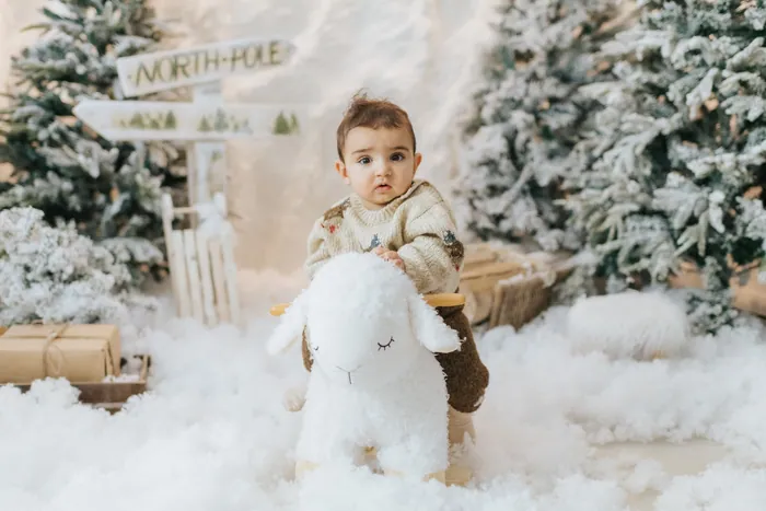 A baby in a cream knitted jumper sitting on a toy sheep in the snow, looking straight at the camera beside a North Pole sign