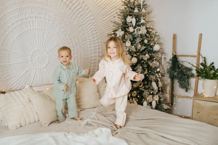 Two toddlers in cream and pale green pyjamas bouncing on a bed in front of a Christmas tree, both grinning