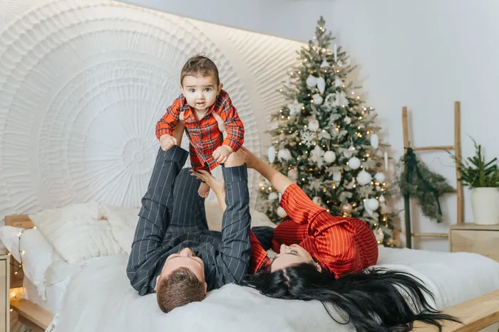 A mother and father lying on a bed lifting their baby in red check pyjamas up above them, a Christmas tree glowing behind