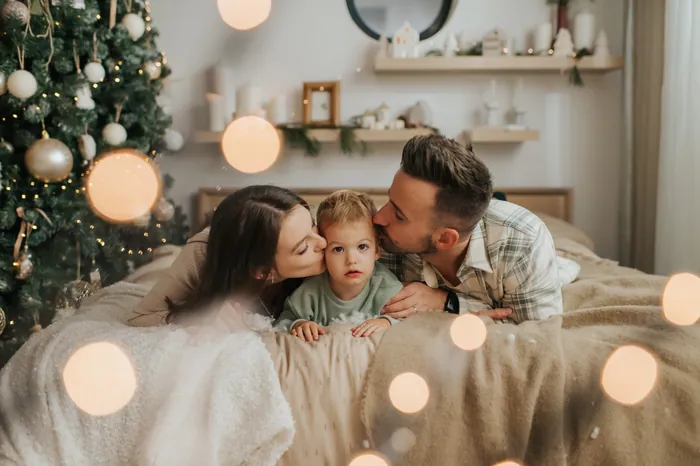 A mother and father kissing their toddler on both cheeks as they lie across a bed, warm Christmas lights in the foreground