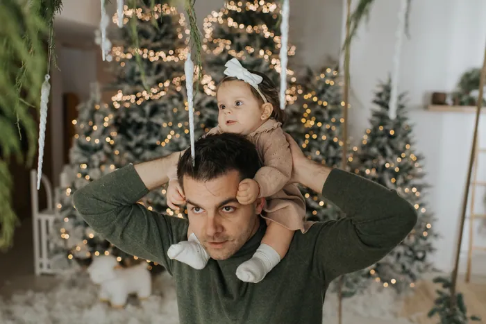 A father pulling a face with his baby daughter on his shoulders, her hands over his ears, flocked trees and lights behind