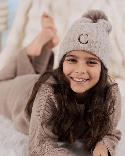 A smiling girl in a knitted hat and soft loungewear lying on a fluffy rug