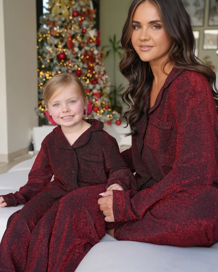 A mother and daughter in matching deep red pyjamas with black piping, a Christmas tree behind them