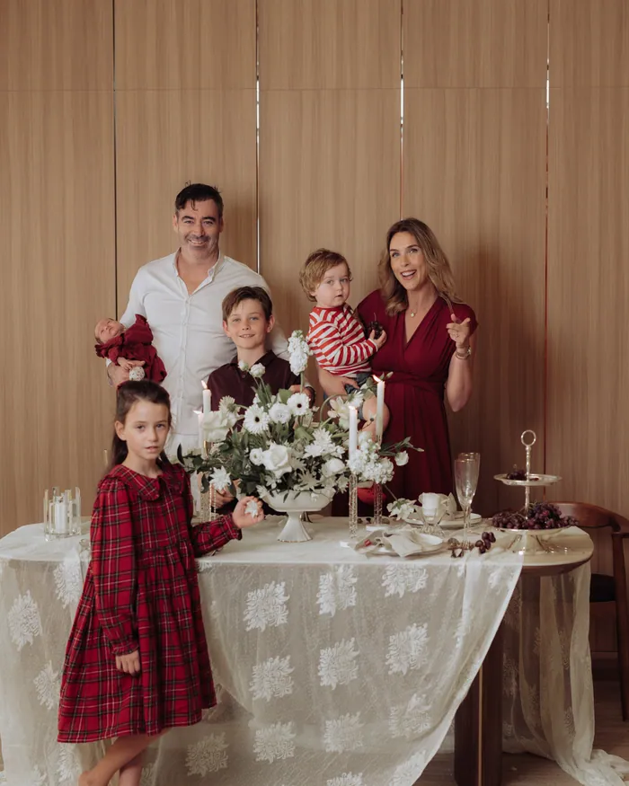 A family of five standing around a lace-covered table set for Christmas tea, the youngest in a red tartan dress