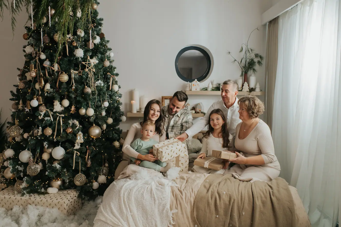 Grandparents, parents and three children opening presents together on a bed beside a tall decorated Christmas tree