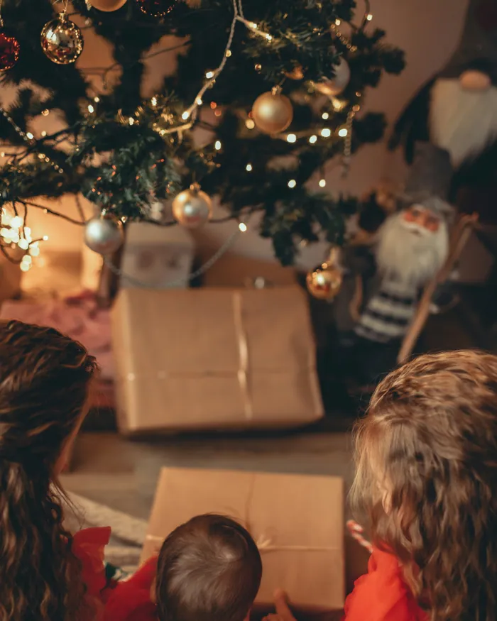 Children in red opening parcels wrapped in brown paper under a lit Christmas tree