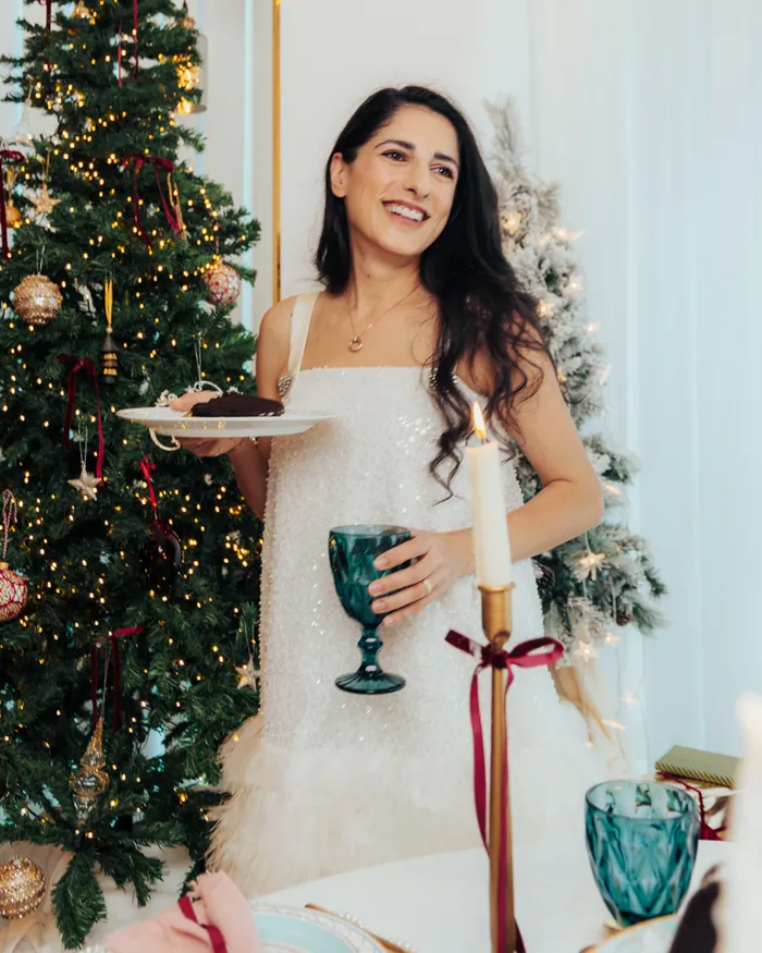 A woman in an ivory dress holding a plate and a teal goblet beside a Christmas tree and a laid table
