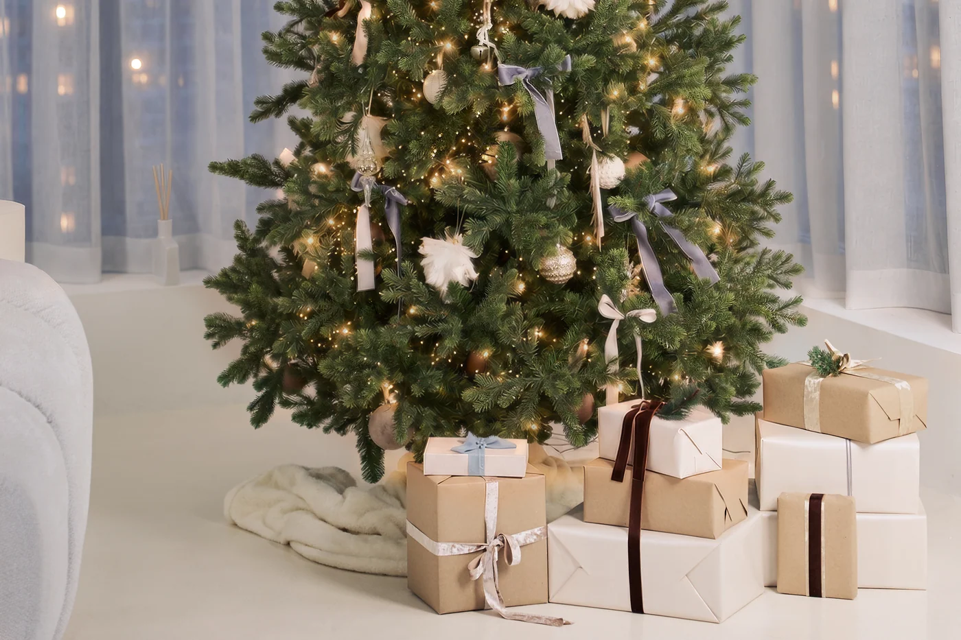 A decorated Christmas tree with white feathers and grey ribbons, wrapped gifts beneath it, in a white room with city windows