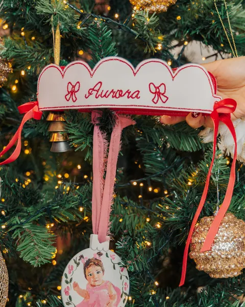 A scalloped white banner embroidered with a name above a painted portrait ornament on a lit Christmas tree