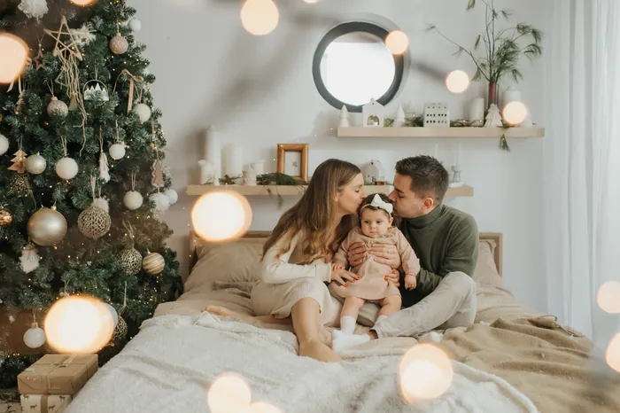 A mother and father kissing their baby daughter on a bed beside a Christmas tree hung with gold baubles